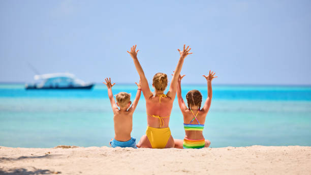 Mother with children on tropical seashore Back view of woman with daughter and son in colorful swimsuits raising arms while sitting on sandy beach of exotic ocean shore sunbathing photos stock pictures, royalty-free photos & images