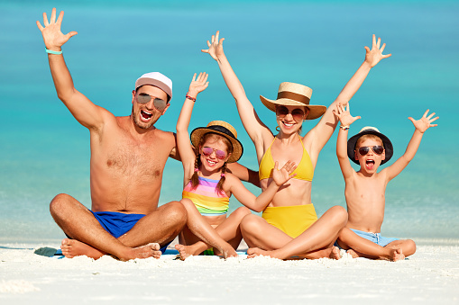 Full length happy family of parents with boy and girl wearing colorful swimsuits and hats with sunglasses raising arms and looking at camera chilling on beach
