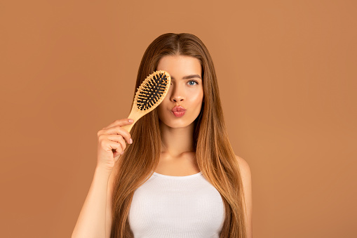 Cosméticos naturales para cabello sedoso. Retrato de una joven encantadora sosteniendo cepillo de pelo cerca de su ojo sobre fondo marrón photo