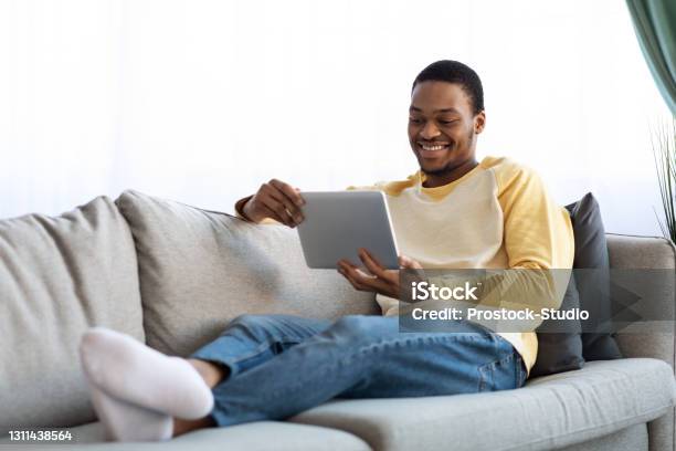 Positive black man reclining on sofa, using digital tablet
