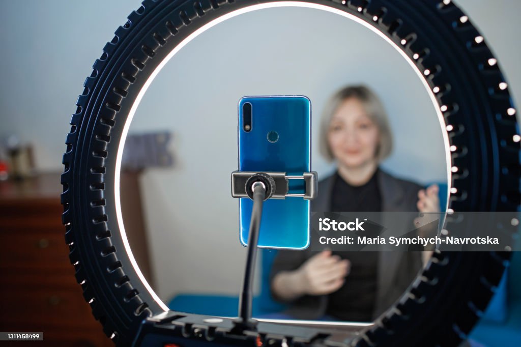 Woman talking to camera and recording video with smartphone and ring light lamp at home, vlogger Video blogging. Business style woman talking to camera and recording video with smartphone and ring light lamp at home, working from home concept, social distant reality Selfie Stock Photo