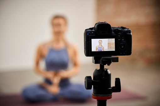 Close-up of camera on tripod filming fitness instructor. Female influencer is vlogging in exercise room. She is at home.