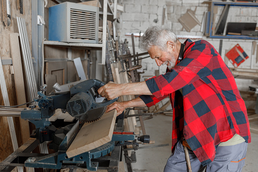 An Elderly Carpenter With Protective Helmet is Manufacturing Wooden Products Using an Electric Saw in Domestic Workshop.