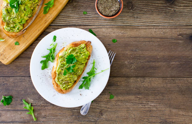 Avocado toast. Shot from above. Avocado toast on a white plate on a rustic wooden table. 
The avocado is sprinkled with chopped parsley and black pepper, and served with fresh arugula. 
Space for copy on the right of the image. 
Stock photo. toasted bread stock pictures, royalty-free photos & images