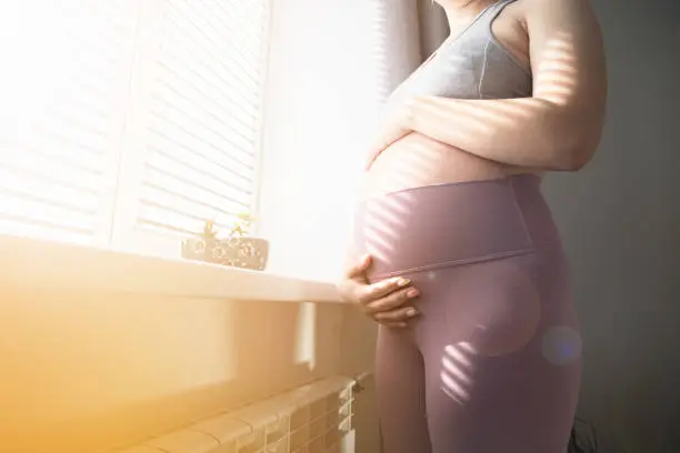 Pregnant woman stands by the window in the room and holds her stomach