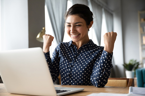 Overjoyed indian ethnicity girl sit at desk looks at laptop screen read incredible news clench fists makes yes gesture celebrate on-line lottery gambling win, getting new job offer feels happy concept