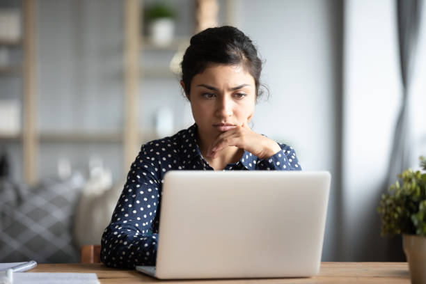 Serious frowning indian woman read email on laptop feels concerned Serious frowning indian ethnicity woman sit at workplace desk looks at laptop screen read e-mail feels concerned. Bored unmotivated tired employee, problems difficulties with app understanding concept worried stock pictures, royalty-free photos & images