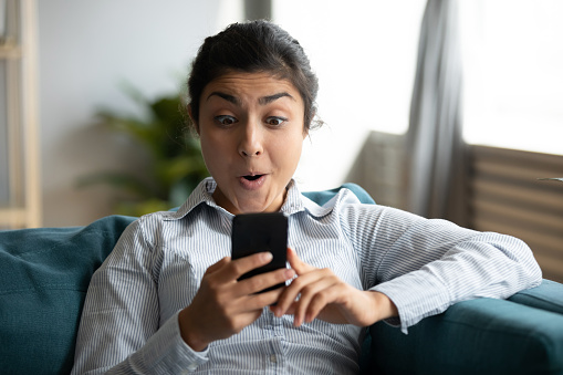 Young indian woman sit on couch holding smartphone with mouth open