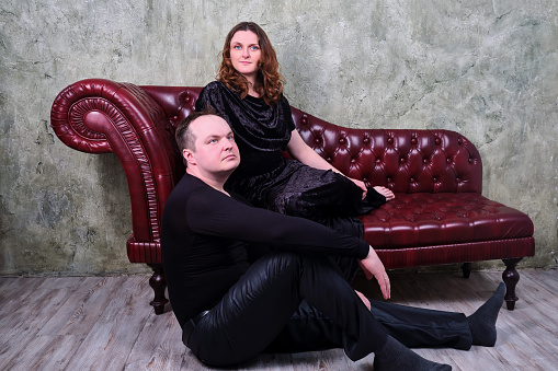 Portrait of a woman and a man seated on a vintage red leather sofa. Studio portrait of an adult couple in retro style