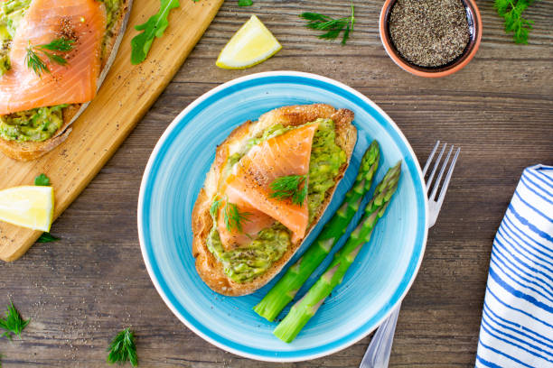 Avocado toast with smoked salmon, shot from above on a turquoise plate on a rustic wooden table. \nThe avocado toast is served with poached asparagus and sprinkled with dill and black pepper. \nStock photo.