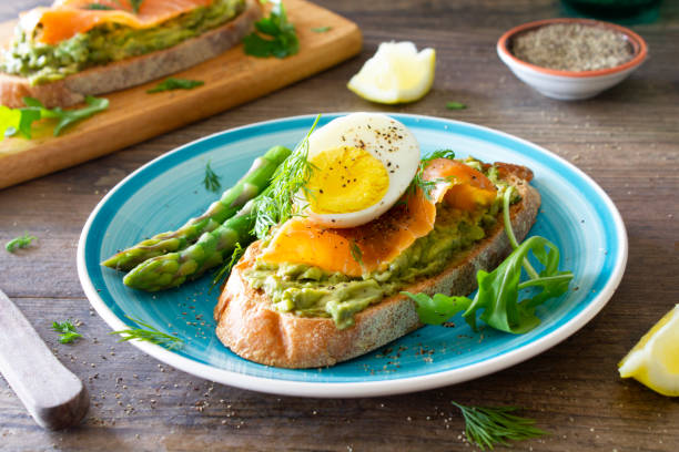 Avocado toast with smoked salmon and egg, on a turquoise plate on a rustic wooden table. \nServed with poached asparagus, fresh arugula and sprinkled with dill and black pepper. \nStock photo.