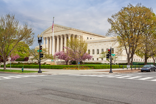 U.S. Supreme Court Building as seen from the First Street, Washington DC, USA. Cloudy Dramatic Sky is in background. Landscaped green bushes, flowers, parked car, traffic lights and red blooming trees are in foreground.\n.