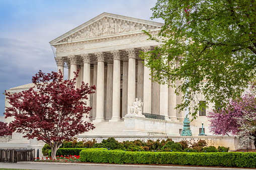U.S. Supreme Court Building as seen from the First Street, Washington DC, USA. Cloudy Dramatic Sky is in background. Landscaped green bushes, flowers and red blooming trees are in foreground.\n.