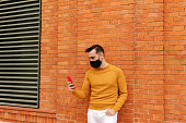 Happy young man with face mask using smartphone in the city. modern stylish yellow clothing. Medical mask and life style.