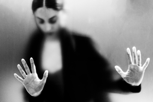 Diffused silhouette of female gesturing fear behind frosted glass