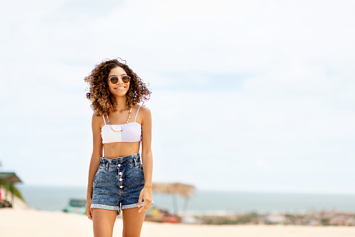 Smiling teenage girl in stylish summer wear walking along a sandy beach during a summer trip to the ocean