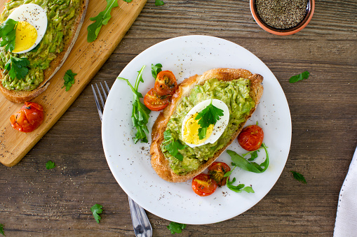 Avocado toast with roasted tomatoes and eggs, shot from above on a white plate on a rustic wooden table. \nServed with fresh arugula and sprinkled with parsley and black pepper. \nStock photo.