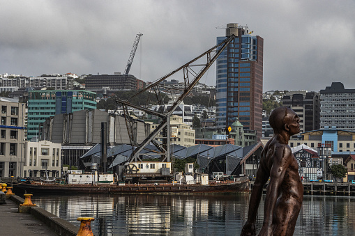 View across the water to the Hikitia, the only working floating crane in Australasia. The boat is anchored on Wellington Waterfront A construction crane is seen in the background amongst the city buildings. In the foreground is a sculpture \