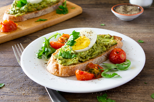 Avocado toast with roasted tomatoes and eggs, on a white plate on a rustic wooden table. 
Served with fresh arugula and sprinkled with parsley and black pepper. Stock photo.