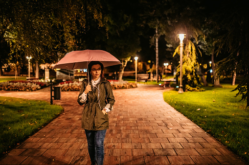 Beautiful young smiling woman walking through public park, listening music on headphones and holding umbrella.