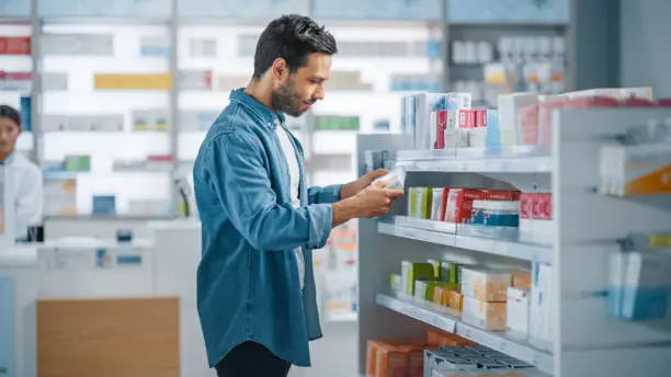 Pharmacy Drugstore: Portrait of Handsome Young Latin Man Searching to Purchase Best Medicine, Chooses between Two Packages of Drugs, Vitamins. Shelves full of Health Care, Wellness, Sport Supplements