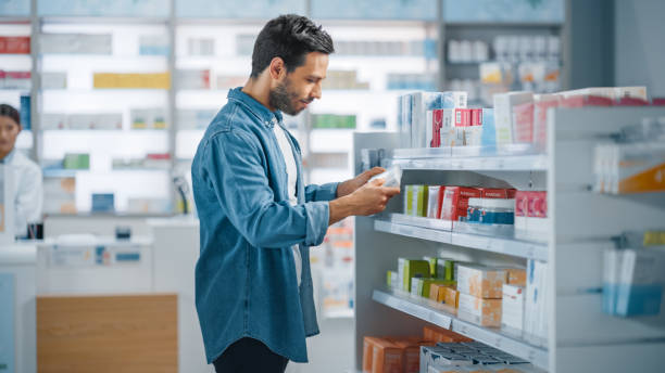 аптека аптека: портрет красивый молодой латинский человек ищет, чтобы купить лучшую медицину, выбирает между двумя пакетами лекарств, вита� - аптека стоковые фото и изображения