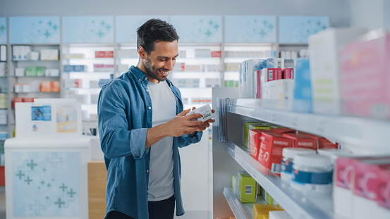 Pharmacy Drugstore: Portrait of Handsome Latin Man Choosing to Buy Medicine Browsing through the Shelf, Successfully finds what he Needs, Smiles Happily. Modern Pharma Store Health Care Products