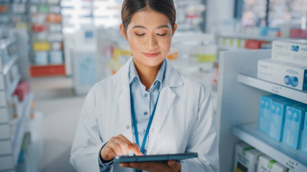 薬局ドラッグストア:美しいアジアの薬剤師の肖像画は、デジタルタブレットコンピュータを使用し、薬のパッケージでいっぱいの彼女の棚の後ろに魅力的に笑顔。中型クローズアップショッ - pharmacy pharmacist smiling pill ストックフォトと画像
