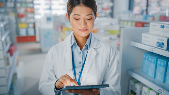 Pharmacy Drugstore: Portrait of Beautiful Asian Pharmacist Uses Digital Tablet Computer and Smiles Charmingly, Behind Her Shelves Full of Medicine Packages. Medium Close-up Shot