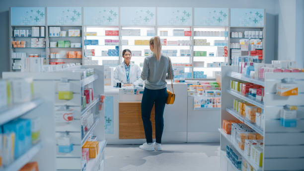 약국 약국: 아름다운 젊은 여자 구입 의학, 마약, 체크 아웃 카운터 옆에 서. 화이트 코트의 여성 계산원은 고객에게 서비스를 제공합니다. 건강 관리 제품이 있는 선반 - 약국 뉴스 사진 이미지