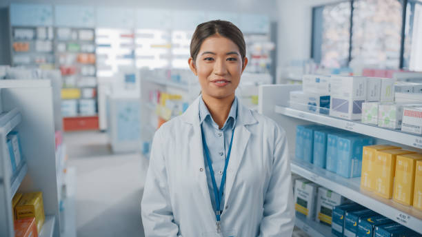 pharmacie pharmacie: portrait de la belle pharmacienne asiatique portant un manteau blanc, regardant l’appareil photo et souriant avec charme, derrière ses étagères pleines de paquets de médicaments. plan rapproché moyen tiré - charmingly photos et images de collection