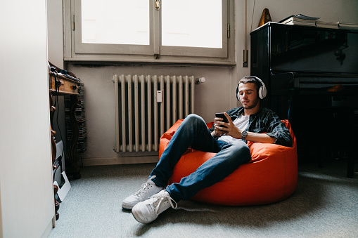 Young adult man texting and listening to music in his bedroom