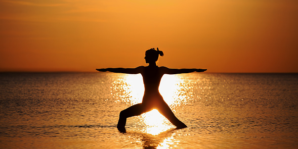 Silhouette of a slender girl practicing yoga at the background of a red-orange sunset on the sea (ocean, lake). Standing position of yoga as exercise - asana  The Warrior (Virabhadrasana). Wide image.