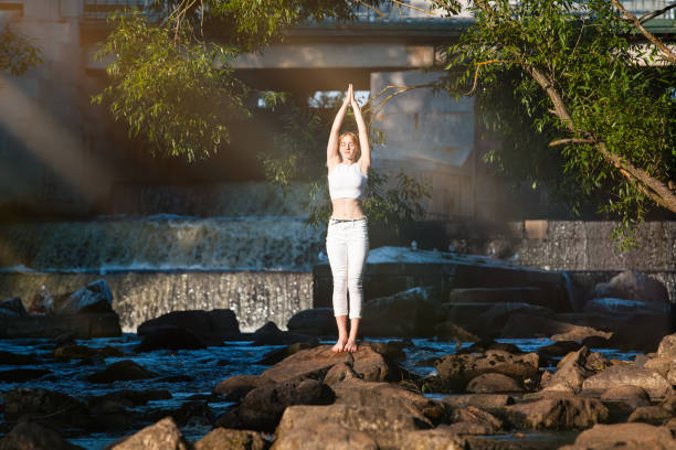 Photo of Young slender woman in white practicing yoga outdoors. Evening meditation and relaxation at the river and waterfall.