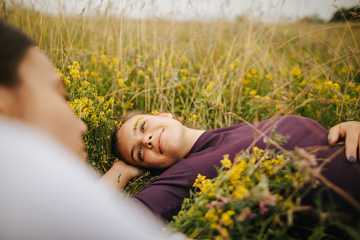 Two young cheerful female friends lie on the green grass among yellow wildflowers and relaxing in beautiful spring nature
