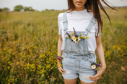 One young woman in white t-shirt and suspenders jeans with wildflowers in top pocket standing on blooming meadow on sunny spring day