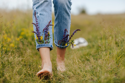 Close-up young female legs walking on green spring grass with wildflowers in legs of denim trousers