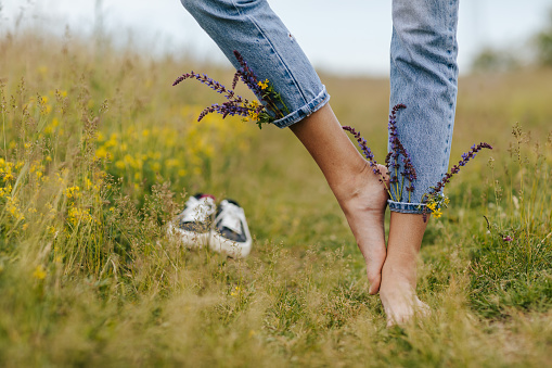 Close-up young female legs walking on green spring grass with wildflowers in legs of denim trousers