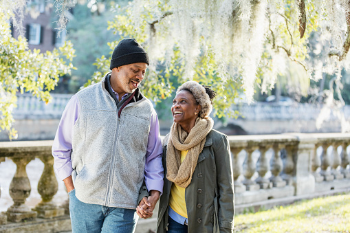 A mature African-American couple taking a walk in the park, along the water, on a sunny fall day. The man is in his 50s, and his wife is a senior woman, in her 60s. They are holding hands, smiling at each other.