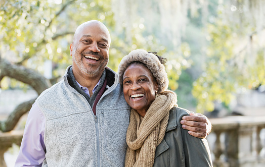 Headshot of a mature African-American couple taking a walk in the park on a sunny fall day. The man is in his 50s, and his wife is a senior, in her 60s. They are standing together, smiling at the camera.