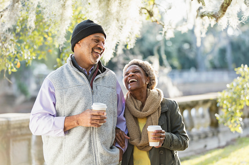 A mature African-American couple at the park on a sunny fall day. The man is in his 50s, and his wife is a senior woman, in her 60s. They are carrying cups of coffee for their morning walk, looking at each other and laughing as they stroll arm in arm.