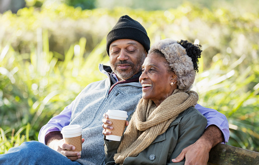 A mature African-American couple hanging out at the park on a sunny fall day. The man is in his 50s, and his wife is a senior woman, in her 60s. They are sitting together on a park bench, relaxing with their morning cup of coffee.