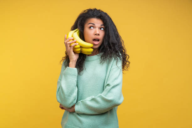 porträt von lustigen positiven lässig gekleidet afro amerikanischen mädchen machen einen anruf über banane telefon isoliert über leuchtenden gelben farbhintergrund hintergrund - banana bunch yellow healthy lifestyle stock-fotos und bilder