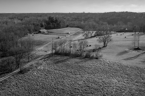 A rural aerial scene by drone in black and white of fields with hay bales, pastures, a barn, and a dirt road leading into the surrounding woods.