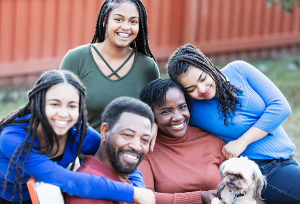 Family with three teenage girls and dog, in backyard An African-American family with three teenage girls together in their backyard, smiling. The parents, in their 50s, are sitting in chairs, and the daughters, 13, 14 and 16 years old, are gathered around them. The mother has the family dog, a shih tzu, on her lap. The focus is on her and the two girls around on her. small florida home stock pictures, royalty-free photos & images
