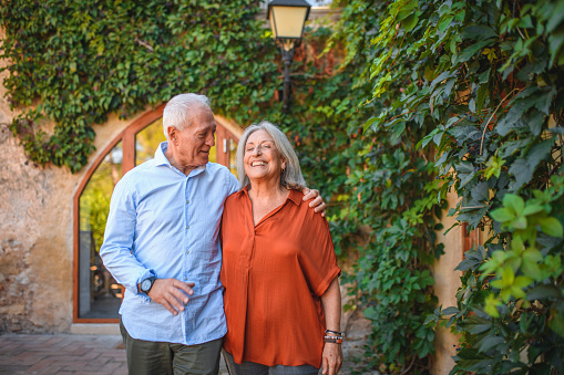 Laughing Spanish seniors with arms around each other walking and talking as they approach camera in outdoor courtyard of family farmhouse.