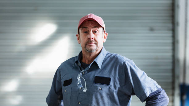 Auto mechanic wearing trucker's hat A mature man in his 50s working as a mechanic in an auto repair shop. He is wearing a trucker's hat, standing with his hands on his hips, looking at the camera with a serious expression. door repairman men manual worker stock pictures, royalty-free photos & images