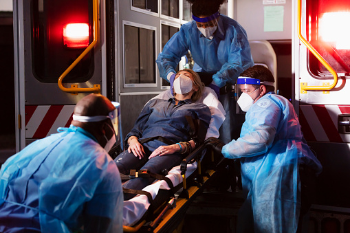 A mature woman in her 40s lying on a stretcher being loaded into an ambulance by three paramedics providing emergency medical care. The EMTs are wearing PPE, including gowns, face shields and masks. They are working during the COVID-19 pandemic, trying to protect themselves from catching and spreading coronavirus.