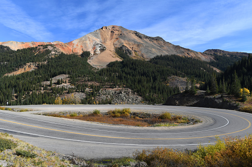 Colorful Red Mountain towers behind famous Million Dollar Highway (U.S. 550) marked with a number of hairpin curves .\nPicture taken in the fall from a roadside above the Uncompahgre Gorge towards the Red Mountain Pass.The original portion of the road was built  in 1883 to connect Ouray and Ironton, The Million Dollar Highway was completed in 1924.