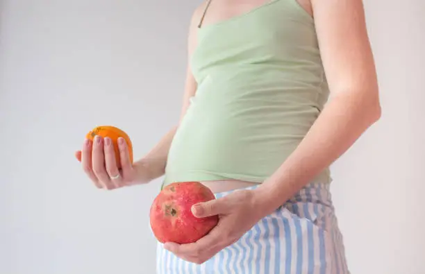 Pregnant girl with fruits. The concept of bearing healthy offspring, replenishing vitamin deficiencies during pregnancy and lactation. Pregnant belly and orange with pomegranate in hands.